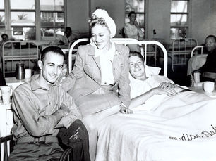 A young woman sits on a hospital bed with a young man in the bed and another sitting in a chair in front of her.