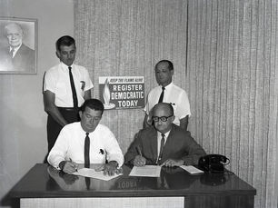 Two men sit at a desk writing with two more men standing behind them. A sign on the wall reads Keep the flame alive register Democratic today.