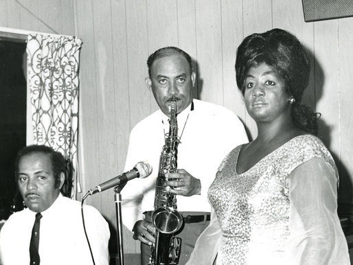 A man sitting at a piano, a man playing saxophone and a woman in front of a microphone.