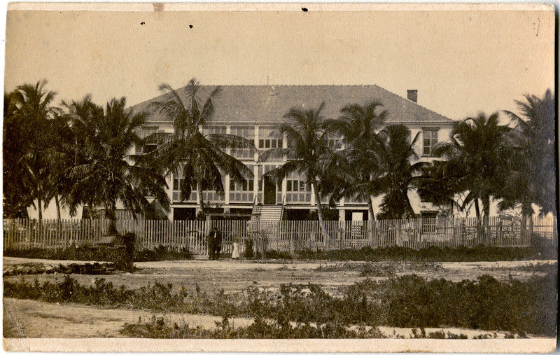 A two story building with a fence, several people and palm trees in front of it.