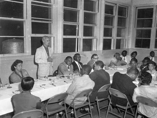 A man standing up addresses a group of people sitting at tables