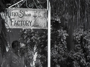 Two people stand under a sign reading kino shoe factory with another sign on the ground reading kindo sandals make an ideal gift.