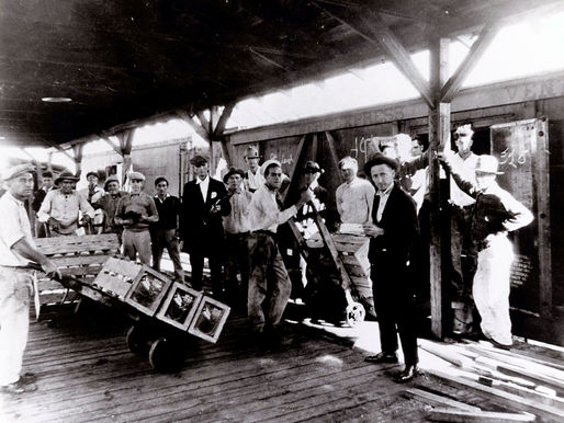 A group of people, two with boxes on dollies, on a platform next to a train car.