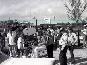 A group of people stand around a movie camera with a sign in the background that reads Cow Key Marina, Key West, Florida