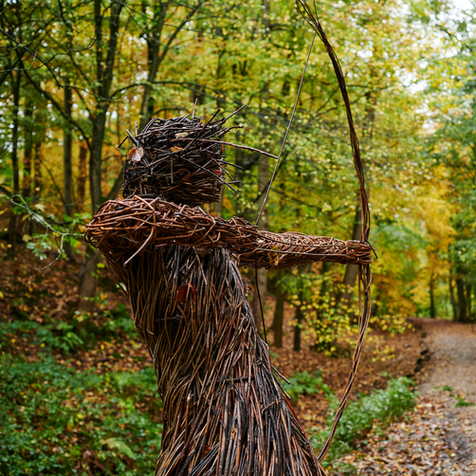 A wicker statue of a female archer aims an arrow down a path through an autumnal wood.