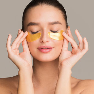 Woman with eyes closed wearing gold under-eye patches, hands on face, against a beige studio background, serene spa mood.