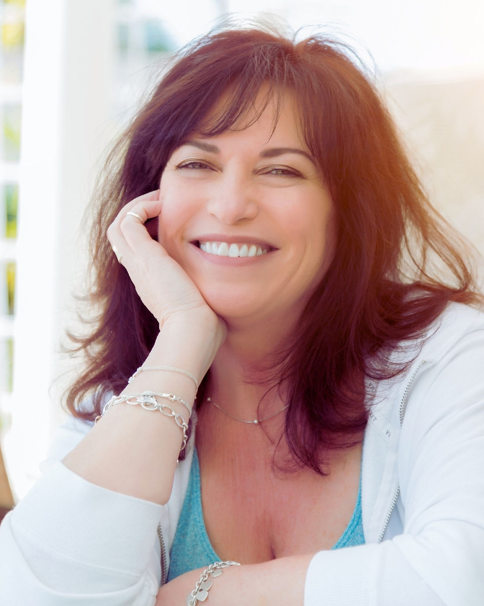 Smiling woman with dark auburn hair rests her chin on her hand in a bright indoor setting, wearing a white jacket and blue top.