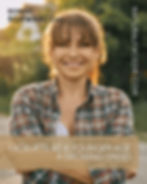 Smiling woman in a plaid shirt stands outdoors at sunset, arms crossed, with blurred trees and a house behind her.