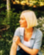 Blonde woman in profile sitting outdoors in a green garden, arms crossed, wearing a light blue shirt, calm and thoughtful.