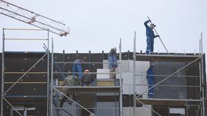 Construction workers on scaffolding install building panels beside a crane on a foggy site under a pale sky.