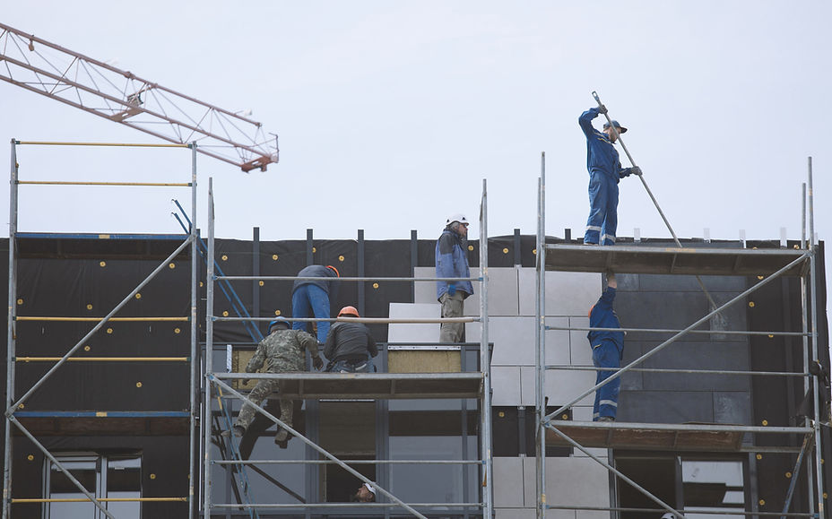 Workers on scaffolding fitting wall panels over dark membrane on a building under construction, with a crane behind