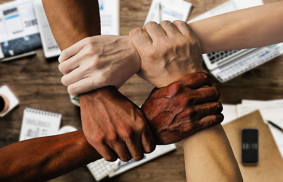 Four diverse hands grasp each other in a unified circle over a wooden desk with papers, phone, and coffee, symbolizing teamwork and solidarity.