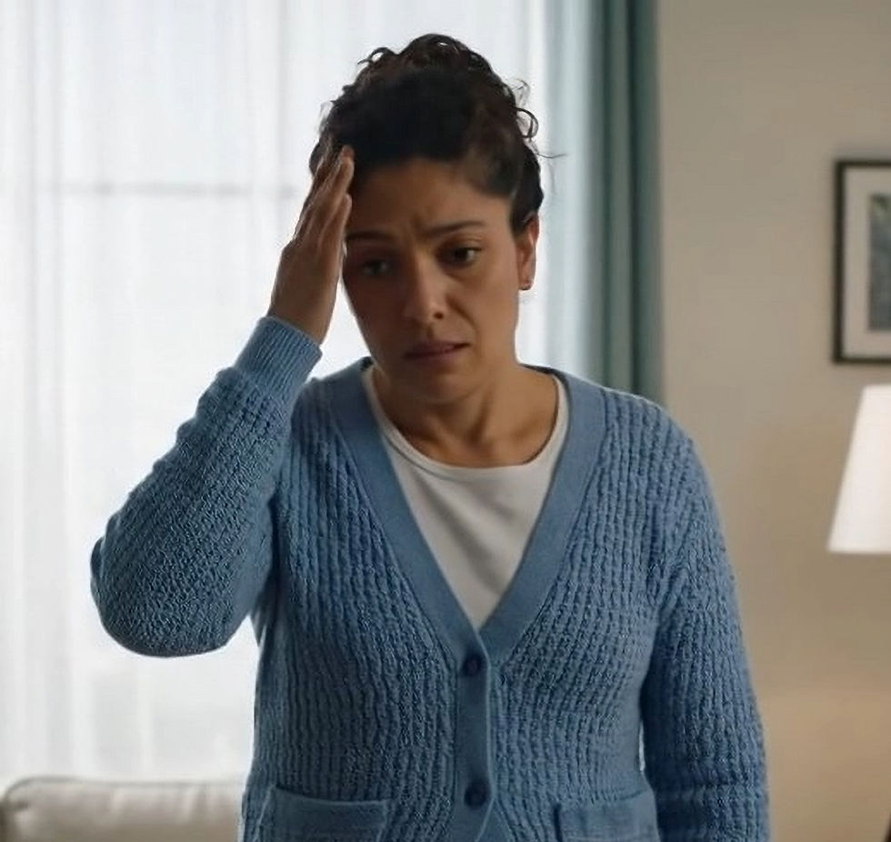 A woman in a blue cardigan holds her head, appearing worried. She stands in a softly lit room with a lamp and framed picture in the background.