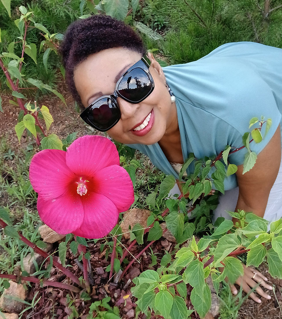 Smiling woman in sunglasses crouches in a lush garden beside a large bright pink hibiscus flower.