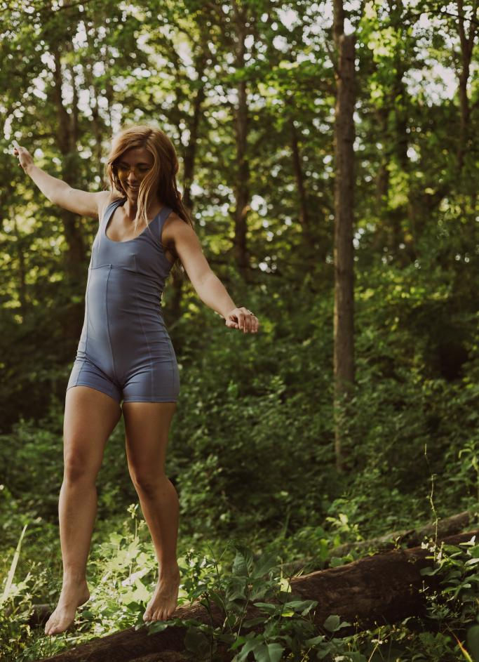 Woman in blue romper balancing barefoot on a log in forest.