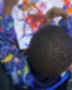 Child painting with a brush on paper, wearing a colorful dinosaur-patterned smock. The artwork involves vibrant reds, blues, and yellows.