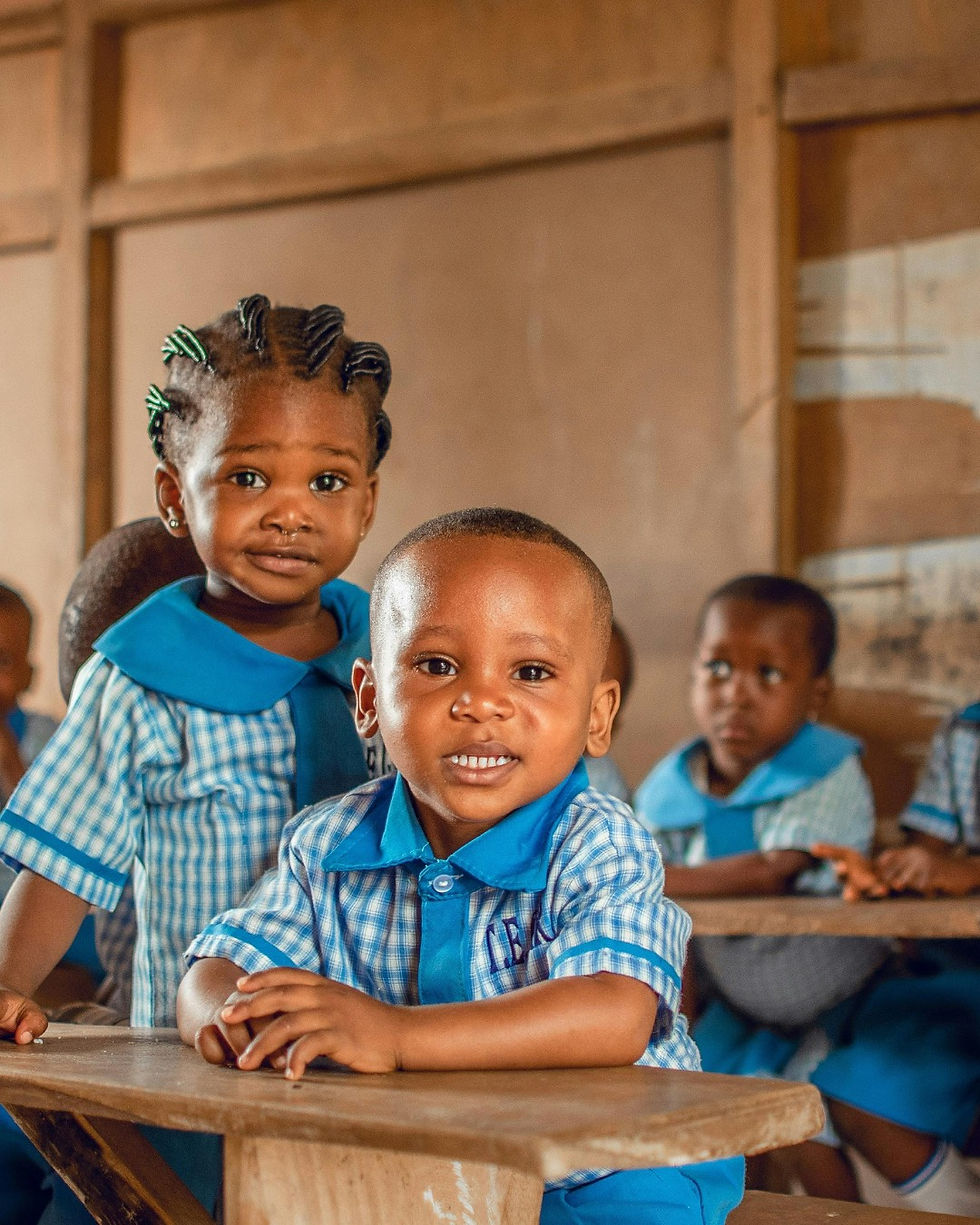 Children in blue checkered uniforms smile at a wooden desk in a classroom.
