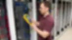 A person in a maroon shirt uses a yellow device to inspect server racks in a data center. Blue and red cables are visible.