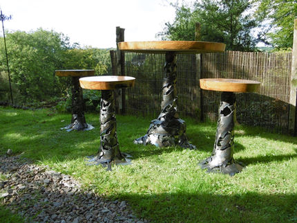 A handcrafted outdoor garden furniture set featuring a high-top table and three bar stools with sculpted steel bases resembling tree trunks and thick, polished live-edge wooden tops on a green lawn.