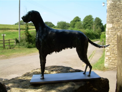 A life-sized handcrafted steel sculpture of a pointer dog with a dark textured finish, standing alert on a metal base atop a stone wall with a countryside road in the background.