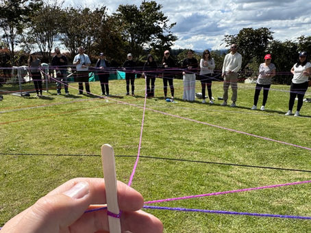 A circle of participants weaves coloured threads across a sunlit field — a regenerative leadership exercise making the living web of stakeholders visible.