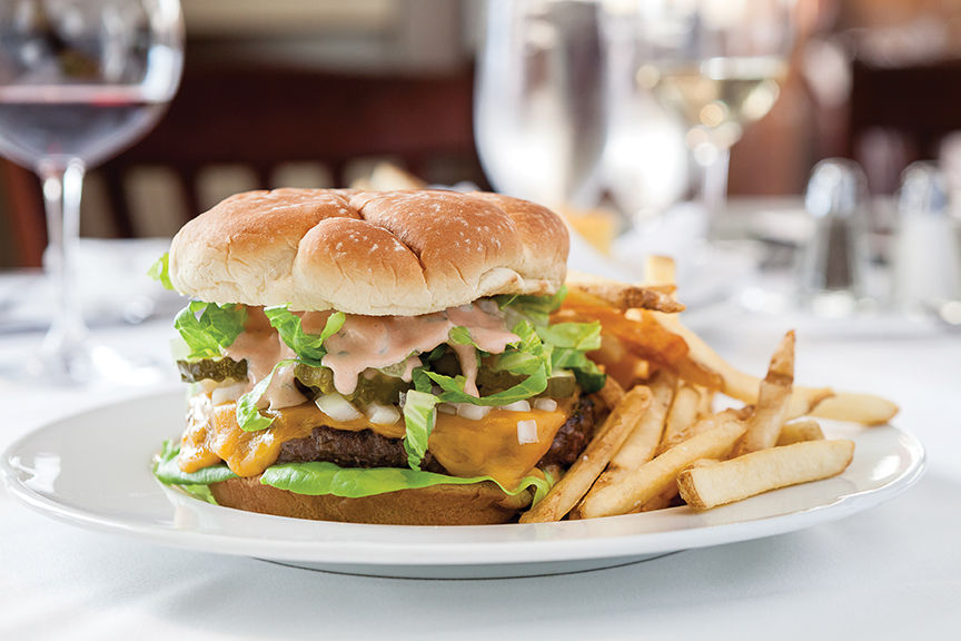 A cheeseburger on a sesame seed bun, topped with lettuce, chopped onions, pickles, and a pink sauce, served with a side of French fries on a white plate