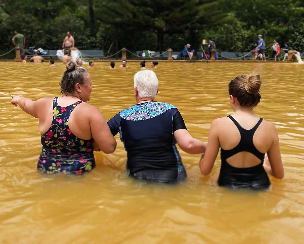 Three people in swimsuits stand in a yellow-brown pool. One points ahead. Greenery and people in the background suggest a relaxed setting.