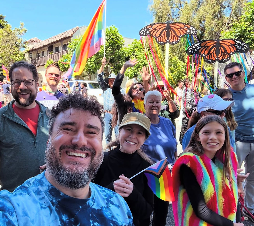A group of progressives at a parade holding rainbow flags and butterfly decorations, smiling under a sunny blue sky with trees and buildings in the background.