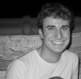 Smiling in a black and white photo, sitting in front of a textured wall. The mood is joyful, with a relaxed atmosphere.