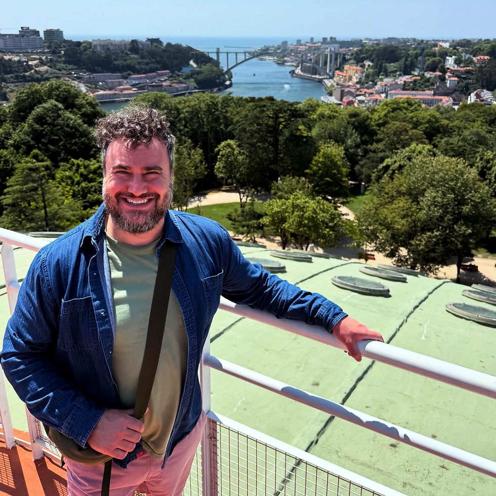 Smiling man in a blue jacket stands on a balcony overlooking a scenic river with a bridge, lush greenery, and a clear blue sky in the background.