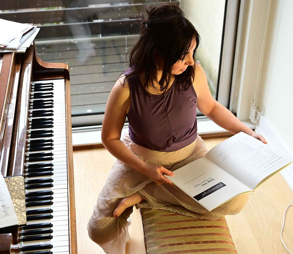 Woman seated beside a piano, reading an open Piano-Yoga book by a bright window, with a calm, focused expression.
