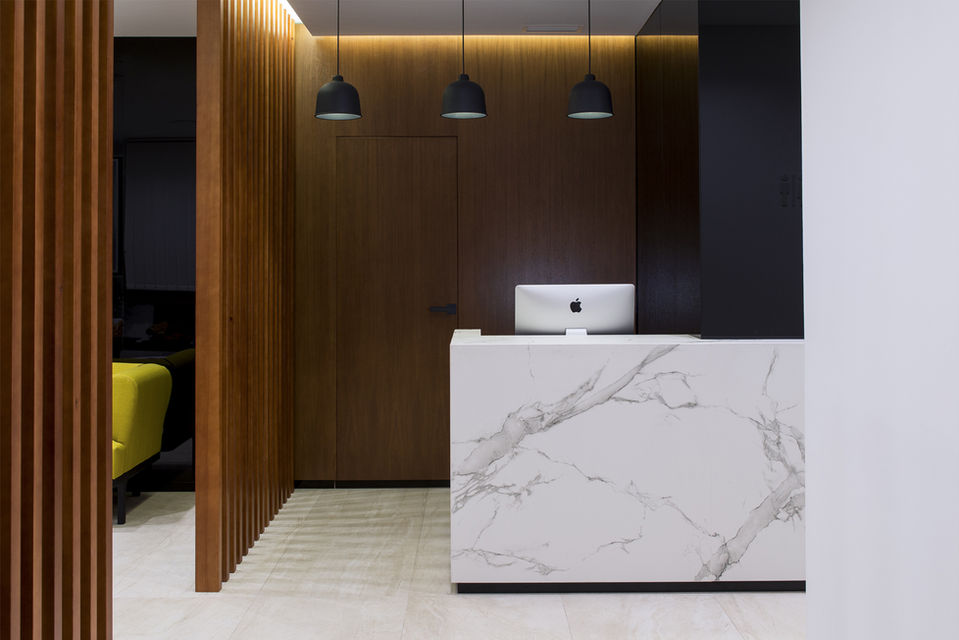 acceso clinica con lamas de madera ,mostrador en piedra blanca con vetas grises y fondo de madera con una puerta oculta.