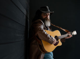 Man in a hat and jacket leans against a dark wooden wall, playing an acoustic guitar. He appears contemplative, set against a dark background.
