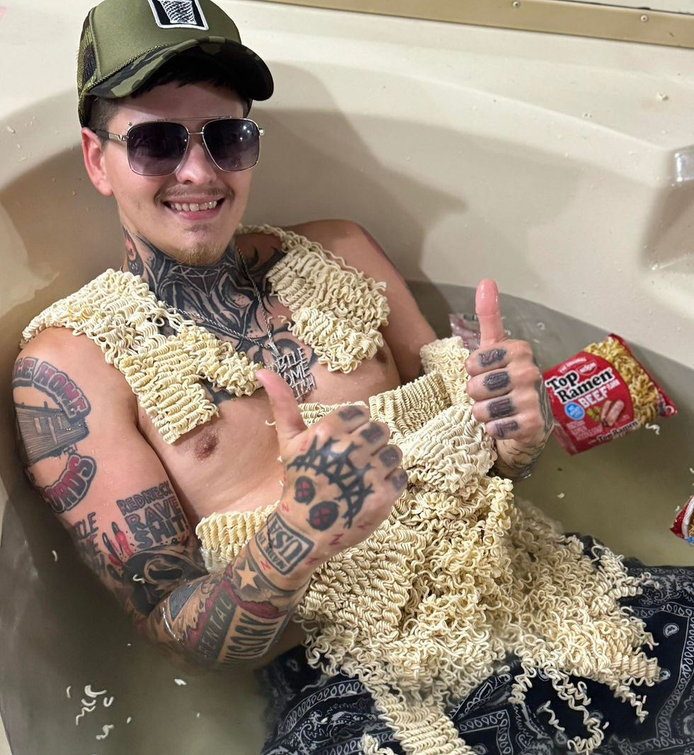 Man in a bathtub wearing ramen packs as clothing, giving thumbs up. Tattoos visible, wearing sunglasses and cap. Pack of Top Ramen nearby.