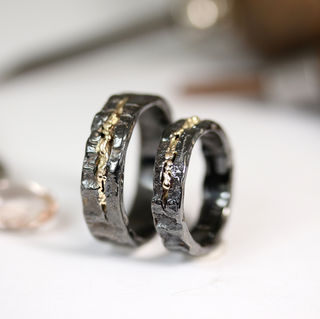 Two handmade wedding rings crafted from black rhodium-coated white gold, featuring textured surfaces and rivers of yellow gold running through the center.