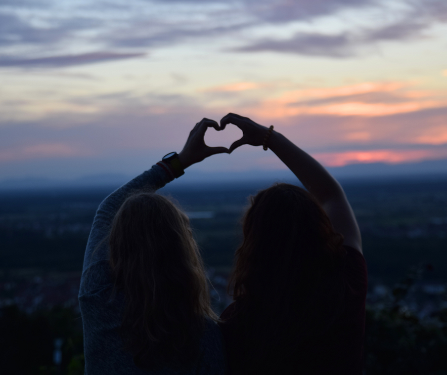 Two women sitting together at sunset, putting their hands together to form a heart shape, symbolizing friendship, connection, and shared joy.