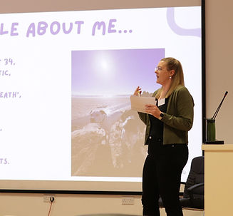 Kate stood on a stage presenting at a conference. The powerpoint is behind her showing half of the slide outlining her personal experiences of being undiagnosed at school.