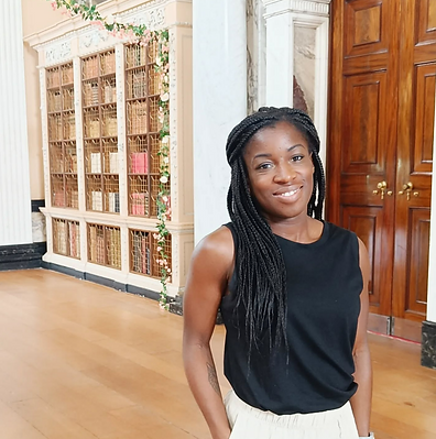 Victoria standing with her hands behind her head in front of a caged library wall to the left and wooden doors to the right. 