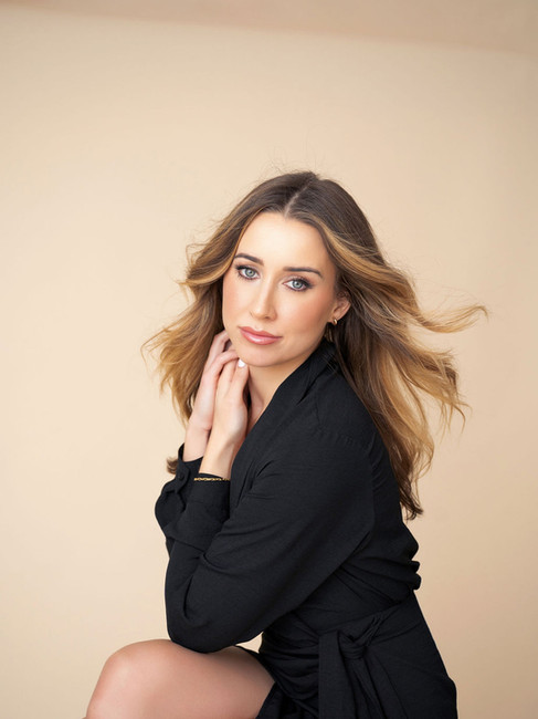 Blonde woman in black blazer outfit sits with crossed legs and gently touches her neck while softly smiling at the camera