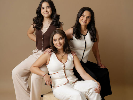 Woman and her two daughters wear brown, black, and white vest outfits and pose together in front of a tan studio backdrop for a mother daughter photoshoot