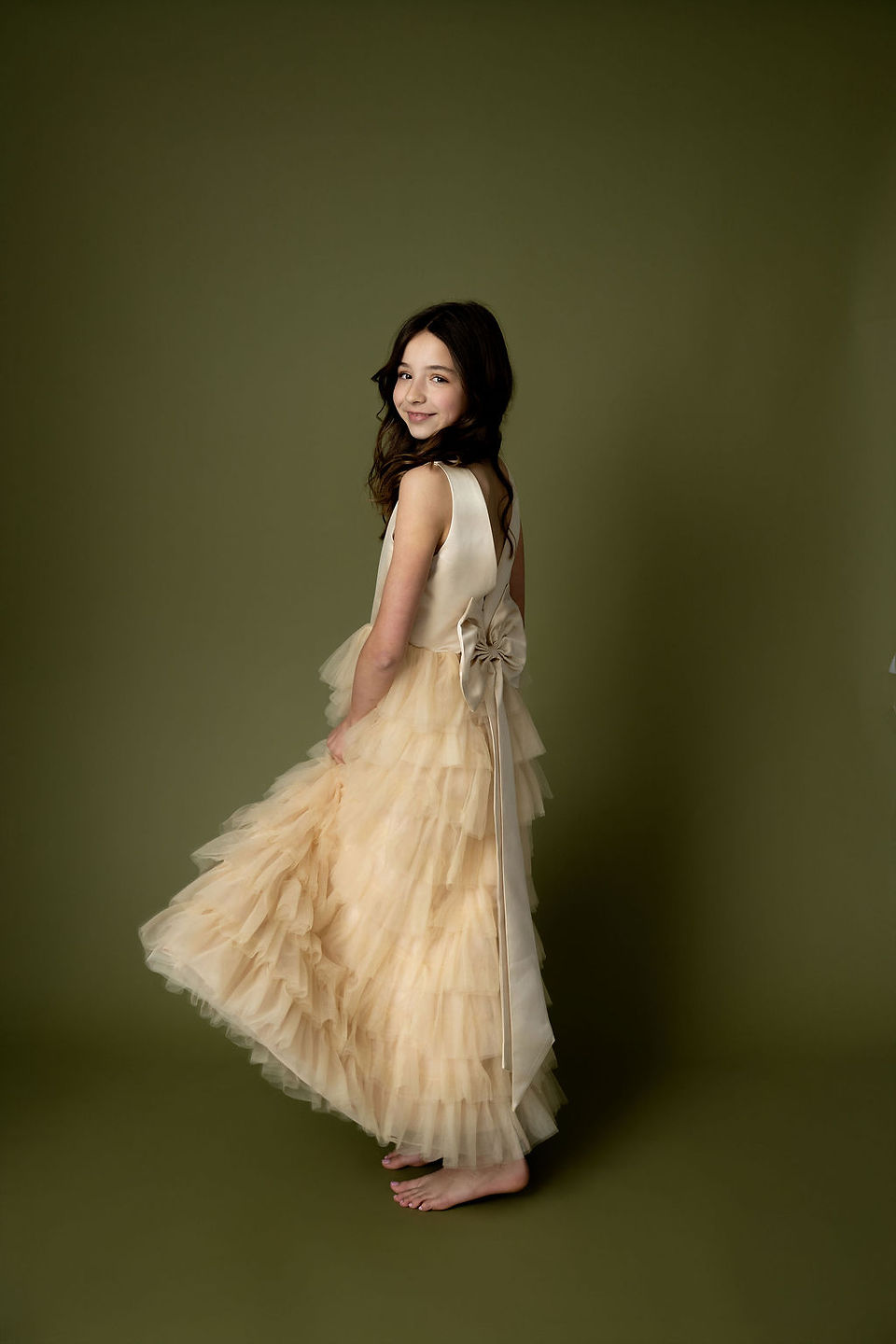 Young girl smiling over her shoulder in a ruffled champagne gown, photographed against an olive green backdrop in a Durham photo shoot studio