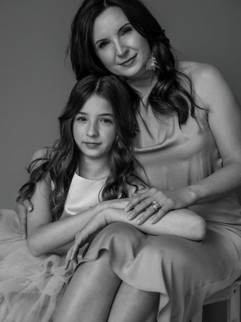 Black and white image of mother and daughter sitting side by side, holding hands and smiling at the camera in an indoor photo studio