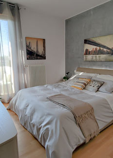 Camera matrimoniale luminosa con letto imbottito, parete effetto cemento, quadri urbani e tende leggere davanti a una portafinestra.
