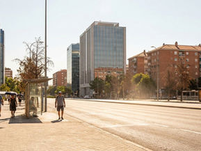 Strada urbana assolata con asfalto, edifici residenziali e uffici esposti al sole, poche zone d’ombra e persone che camminano nel caldo estivo.
