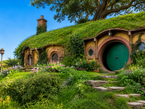Casa Baggins integrata nella collina, con porta e finestre rotonde, tetto erboso, camino, grande albero e giardino fiorito.
