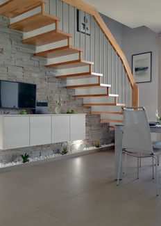 Soggiorno moderno con parete in pietra, scala in legno a sbalzo e arredi minimalisti; tavolo da pranzo con sedie grigie e illuminazione a sospensione.
