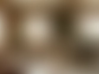 Un soggiorno luminoso in stile biofilico che integra armonicamente le esigenze di adulti e bambini. A destra, un adulto lavora al computer seduto su un divano dalle tinte neutre; a sinistra, un bambino gioca a terra su un grande cuscino, accanto a mobili bassi in legno chiaro proporzionati alla sua altezza. L'ambiente è caratterizzato da grandi finestre, travi a vista in legno, toni caldi e piante da interno.