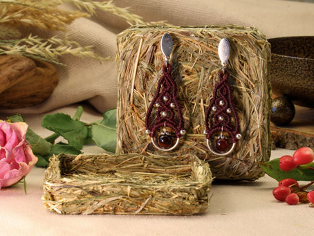 Exklusives Schmuckhandwerk von Aranya’s Art: Handgefertigte Makramee-Ohrringe in Weinrot mit dunklen Edelsteinperlen, präsentiert auf einer nachhaltigen Zero Waste Heuverpackung. Dekoriert mit einer Trockenrose und roten Beeren in einer natürlichen Boho-Ästhetik.