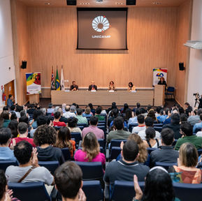 Foto do Auditório Milton Santos do IG - Unicamp. Imagem aberta com o público de costas sentado em cadeiras azuis no primeiro plano. No segundo plano, mais ao fundo, uma mesa com quatro pessoas sentadas e, à direita, um púlpito com uma pessoa de pé. Ao fundo, uma parede bege com uma tela de projetor aberta contendo a imagem do símbolo da Unicamp.