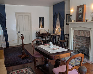 A period-accurate 1860s parlor featuring a small square piano, wooden chairs, and Victorian-style home decor.